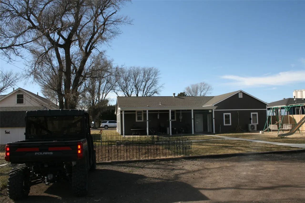 A dark utility vehicle parked on a dirt lot facing a one-story, dark-sided house with a porch under a clear blue sky.