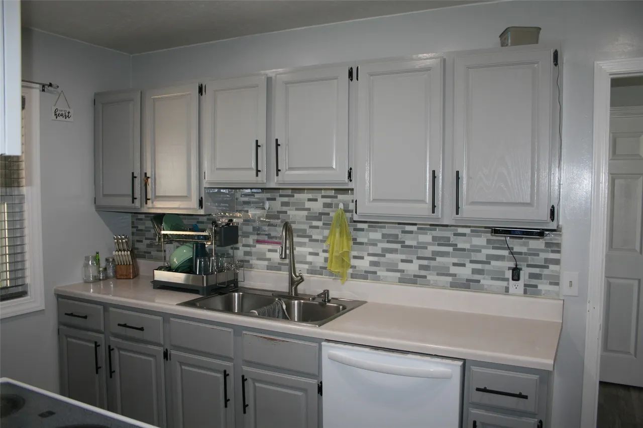 A kitchen with gray cabinets, a tiled backsplash, white countertops, a stainless steel sink, and a white dishwasher.