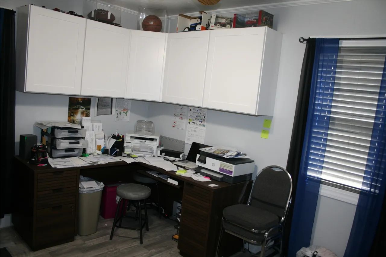 A home office corner with white wall cabinets above an L-shaped desk, a printer, papers, and a gray rolling stool.