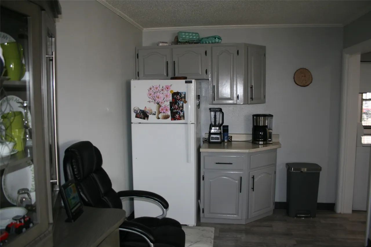 A kitchen area with a white refrigerator, grey cabinets, a blender, a coffee maker, and a black office chair.