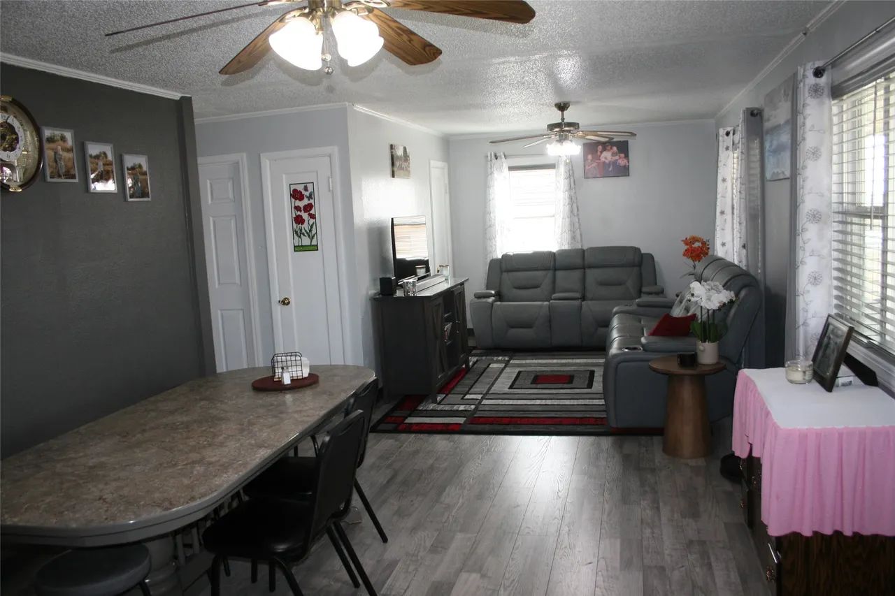 A dining table sits in the foreground of an open-concept living area with grey furniture, wood flooring, and a rug.