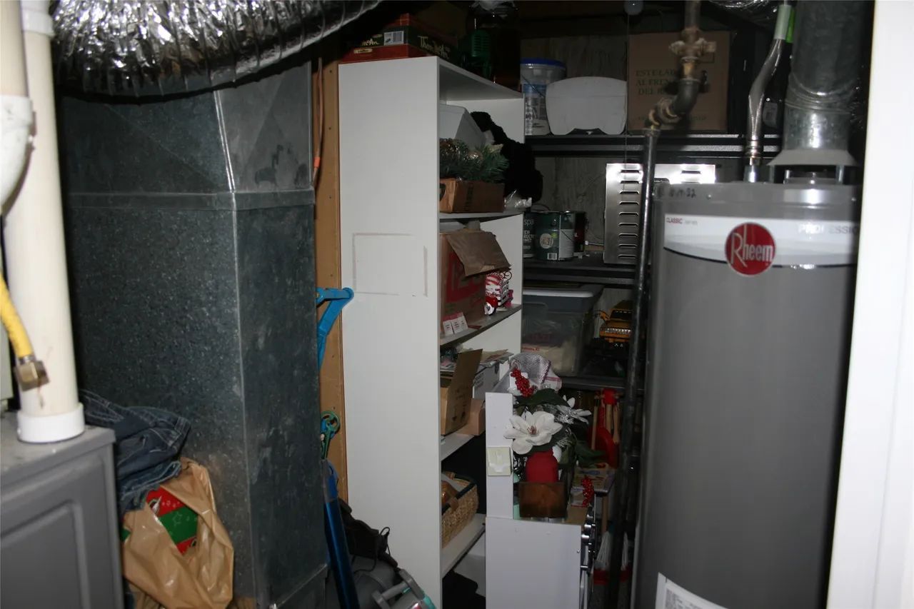 A cluttered utility closet with a water heater, a metal air duct, and a white shelving unit filled with various items.
