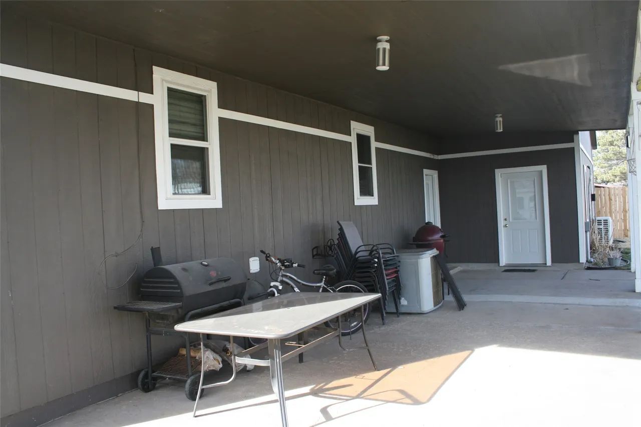 A covered concrete patio with a table, grill, and outdoor chairs against the dark gray wall of a house.