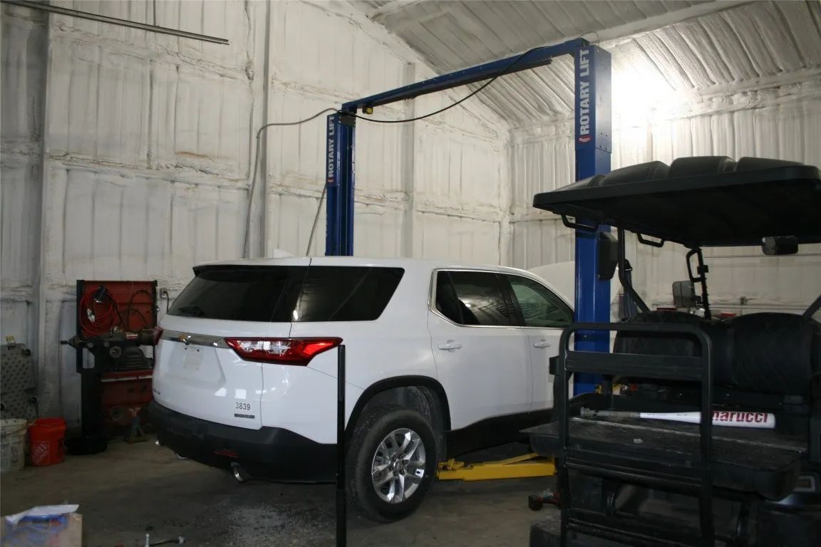 A white SUV parked inside a garage workshop, lifted by a two-post blue automotive lift next to a utility vehicle.