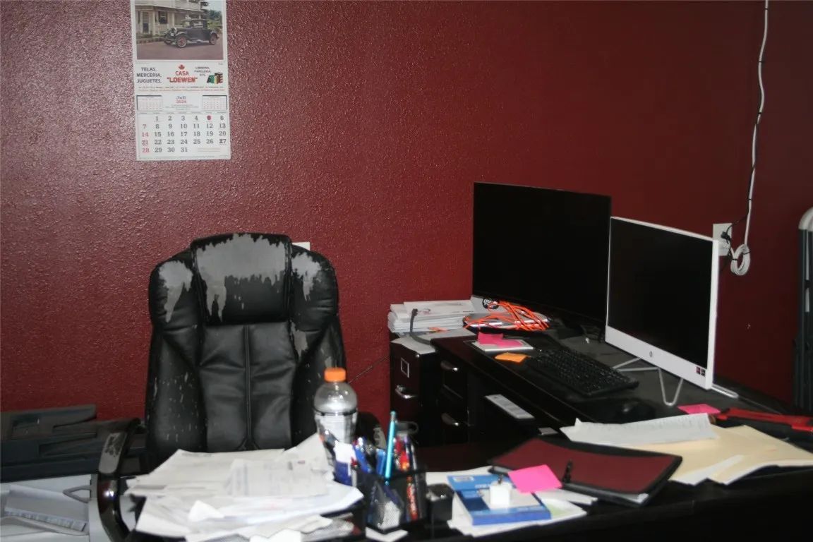 A desk with two computer monitors, a worn office chair, and cluttered papers against a dark red wall in an office.