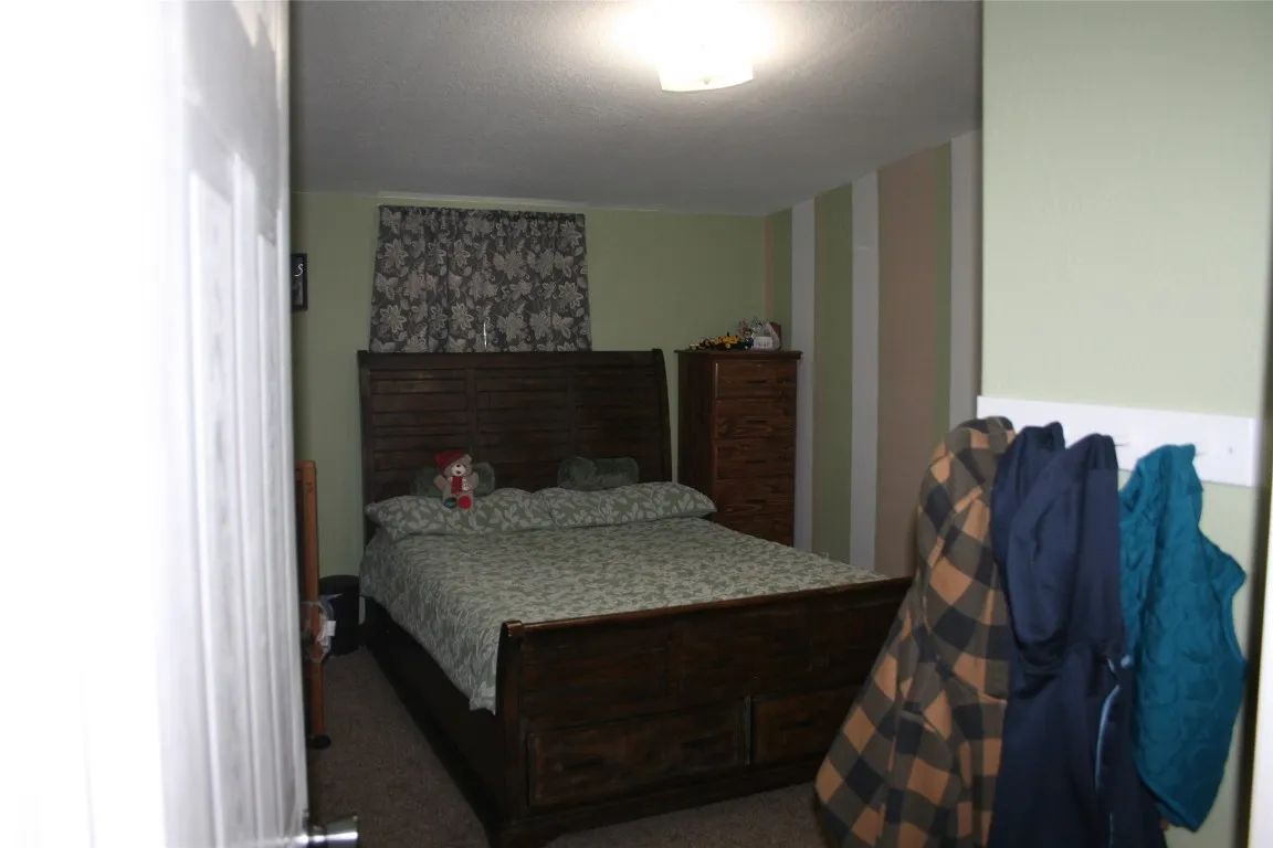 A bedroom view through a doorway, showing a dark wood bed, a matching tall dresser, and coats hanging on a wall hook.