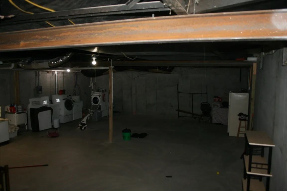 Unfinished basement with exposed ceiling beams, concrete walls, a laundry set, and utility shelves on the right.