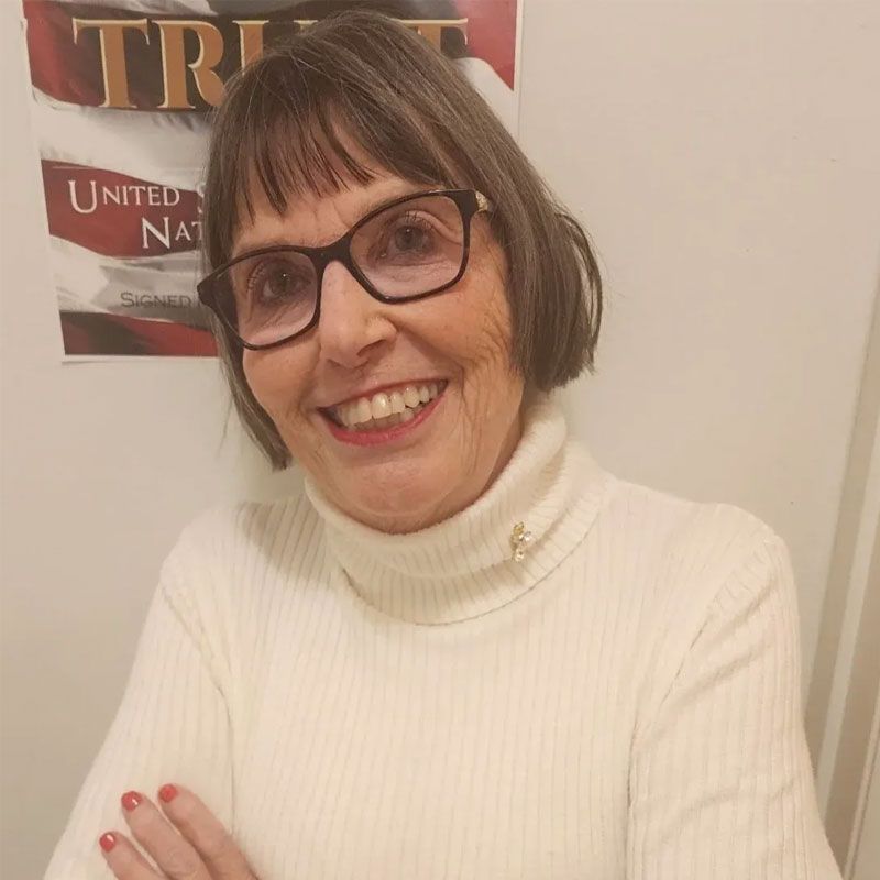 Woman with glasses smiles, wearing a white turtleneck sweater, near an American flag poster.