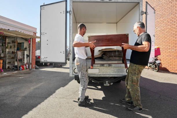 Two uniformed men are transporting boxes into a living room as part of a furniture moving service.
