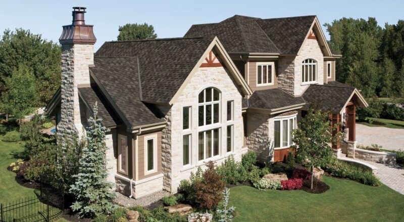 Stone-faced, two-story house with a dark roof, chimney, and well-maintained landscaping in a sunny setting.