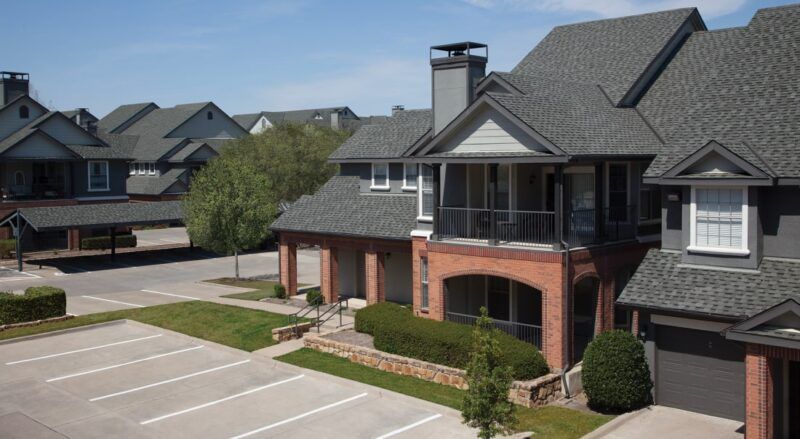 Townhouses with gray roofs and brick accents sit in a sunny neighborhood with a parking lot.