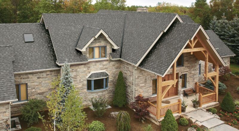 Stone house with a dark gray shingled roof, wooden porch, and landscaping.
