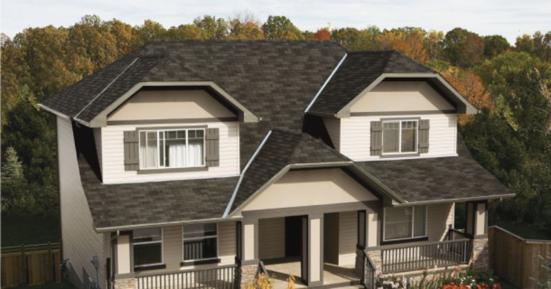 A two-story house with a dark gray roof, tan siding, and white trim. Two dormers and a front porch are visible.
