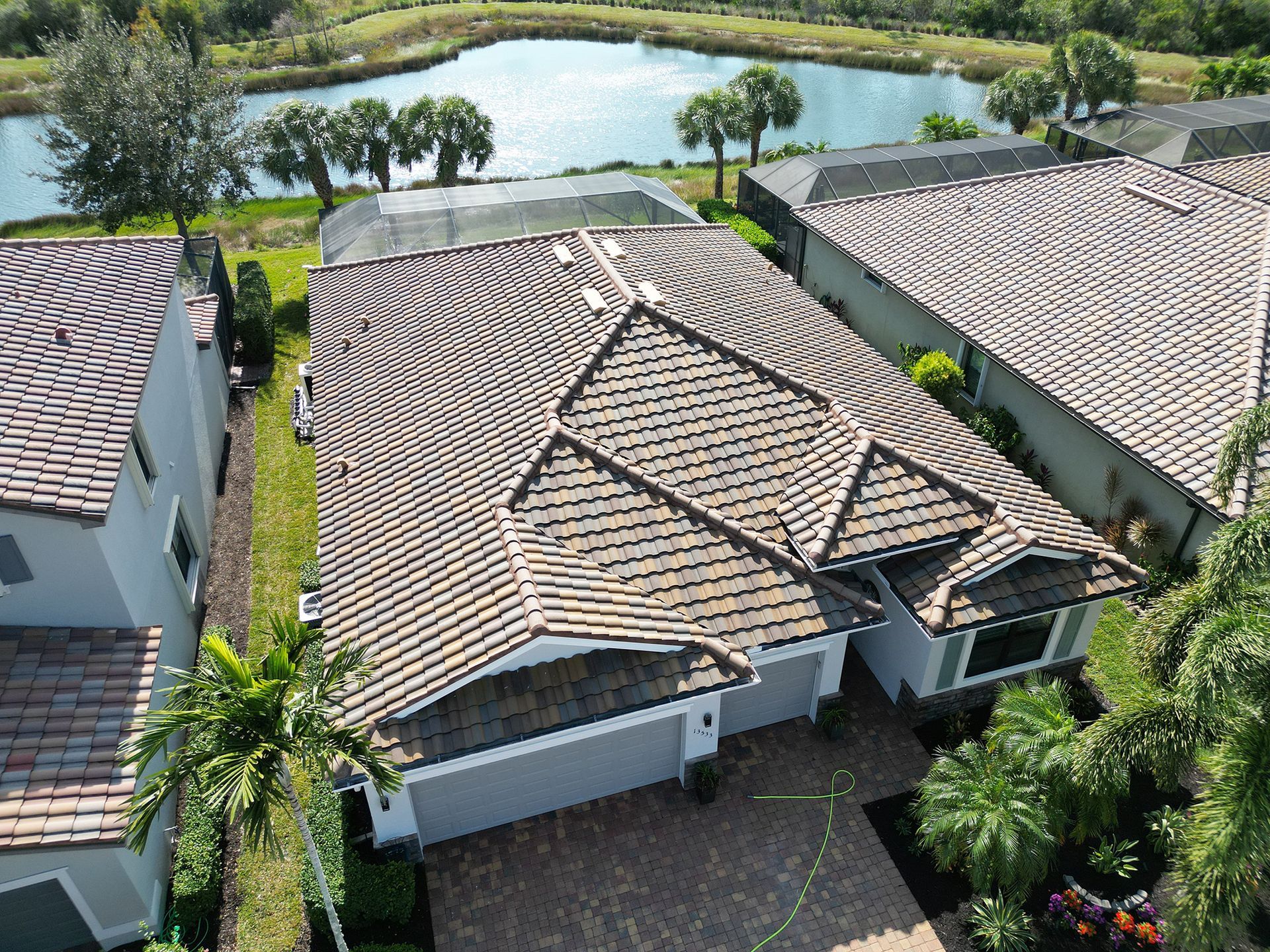 Aerial view of a tiled-roof house with a driveway, palm trees, and a pond behind it