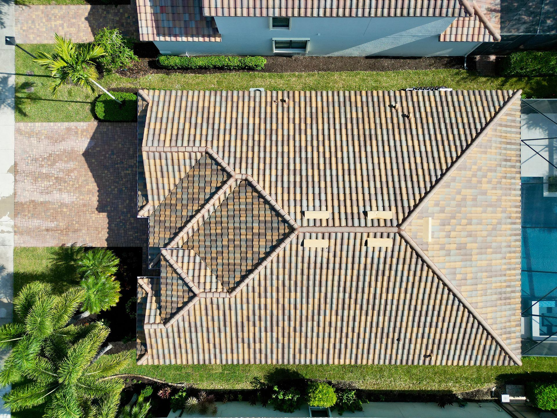 Aerial view of a house with a tan tiled roof, surrounded by green shrubs and a paved driveway.