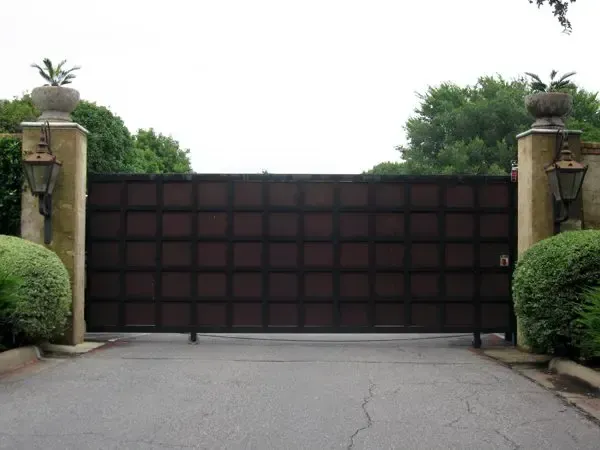 Dark brown, square grid gate at a driveway entrance, flanked by stone pillars with lanterns and greenery.