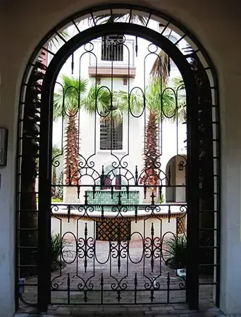 Ornate wrought iron gate framing a courtyard with fountain, palms, and a multi-story building.