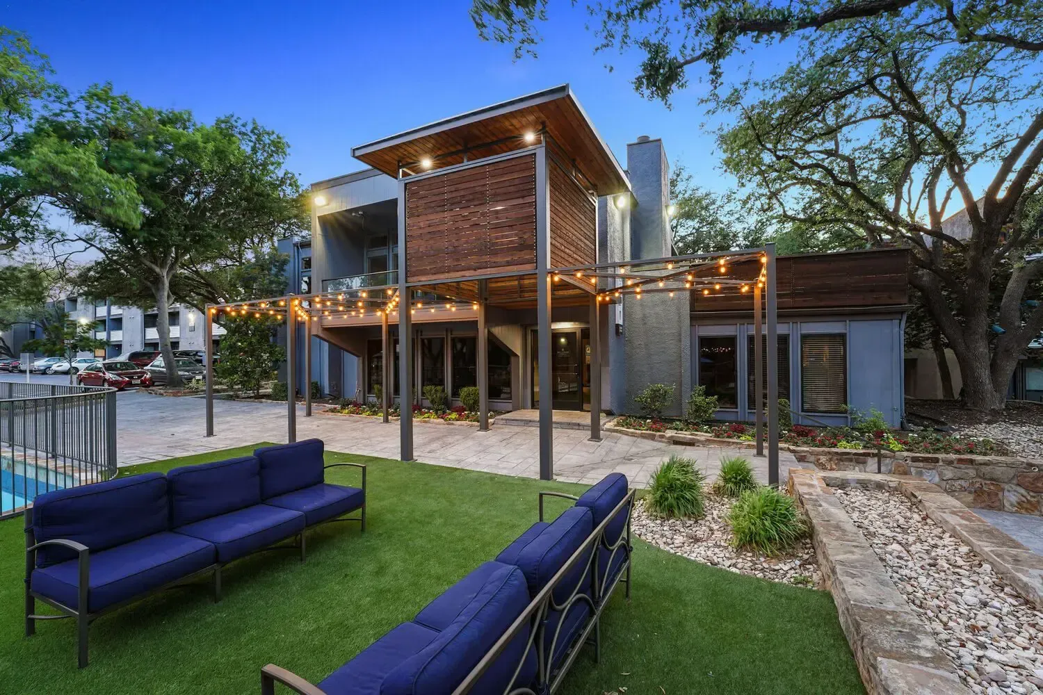 Modern building with blue couches on lawn near a pool and string lights.