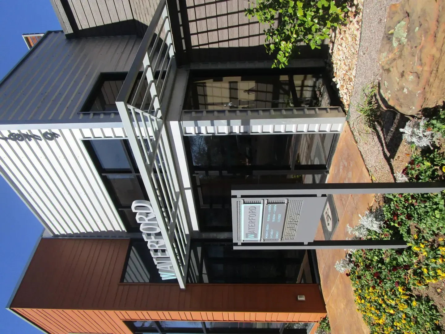 Modern building with a sign, brown and white exterior, blue sky, plants, and sidewalk.