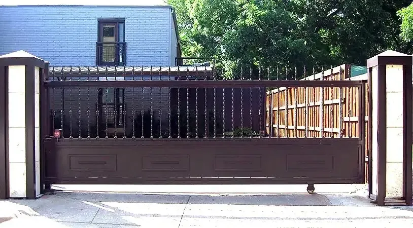 Brown metal sliding driveway gate, vertical bars and solid panels, with ornate pillars, set against a residential house.