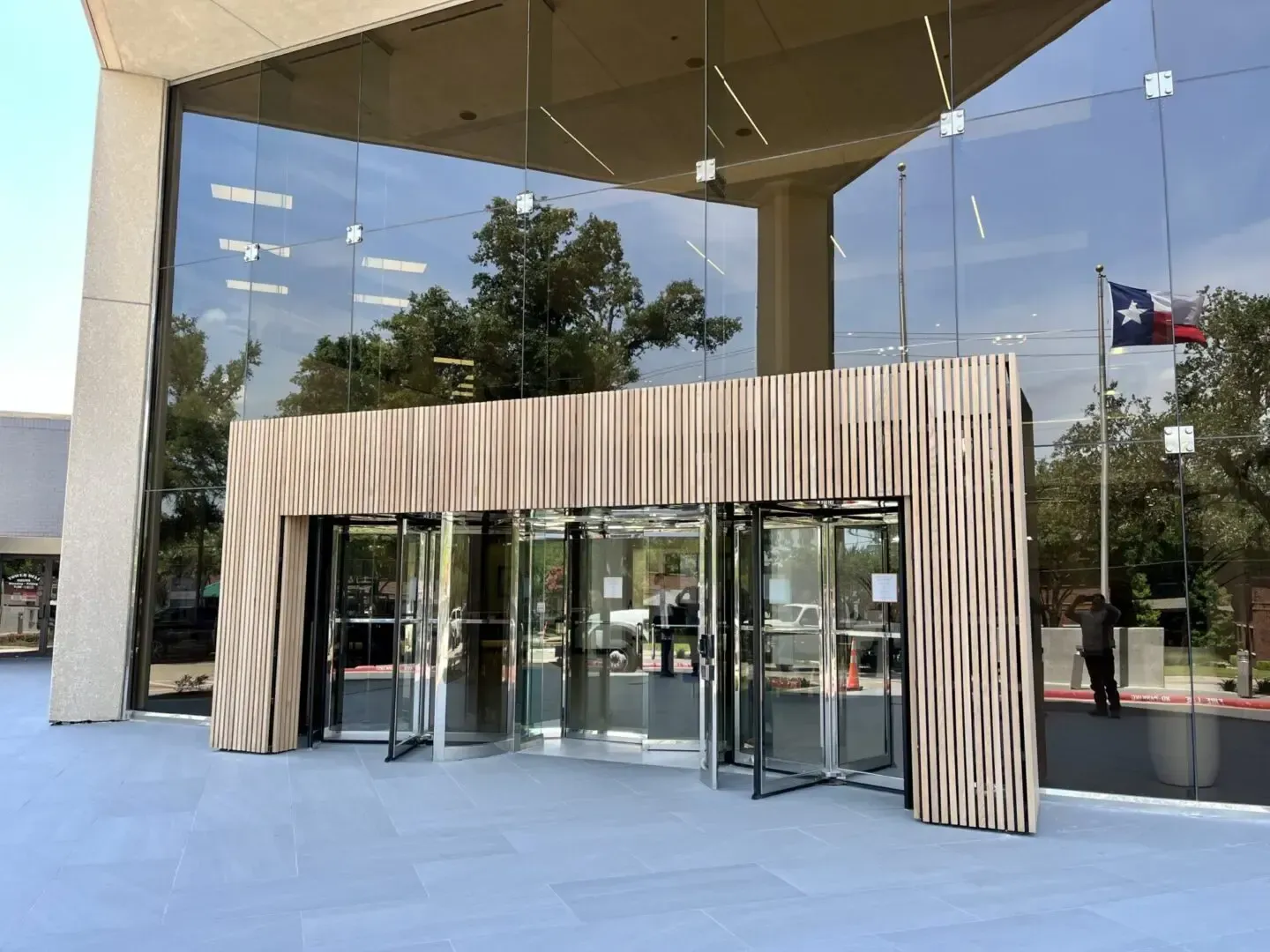 Glass-walled building entrance with revolving doors, wooden frame, and Texan flag on a sunny day.