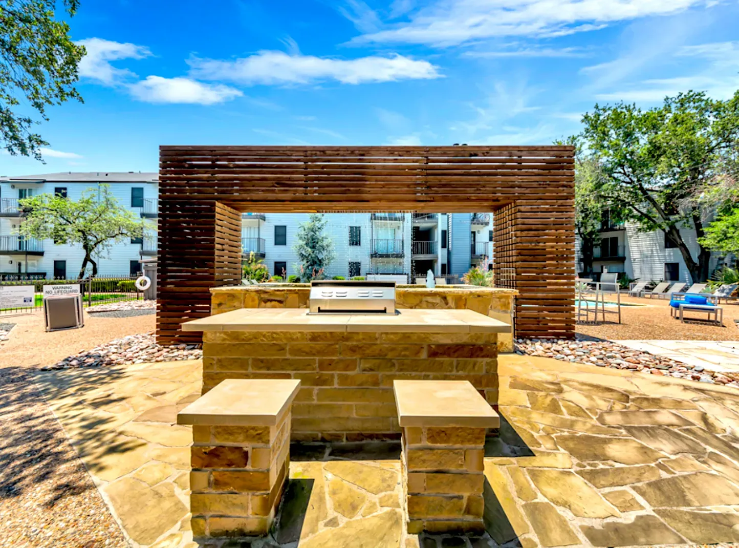 Outdoor grill area with a stone countertop and wooden overhead structure in front of apartment buildings under a blue sky.