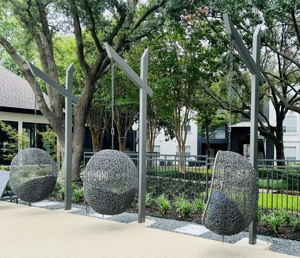 Three hanging spherical chairs of woven material on metal posts in outdoor area.