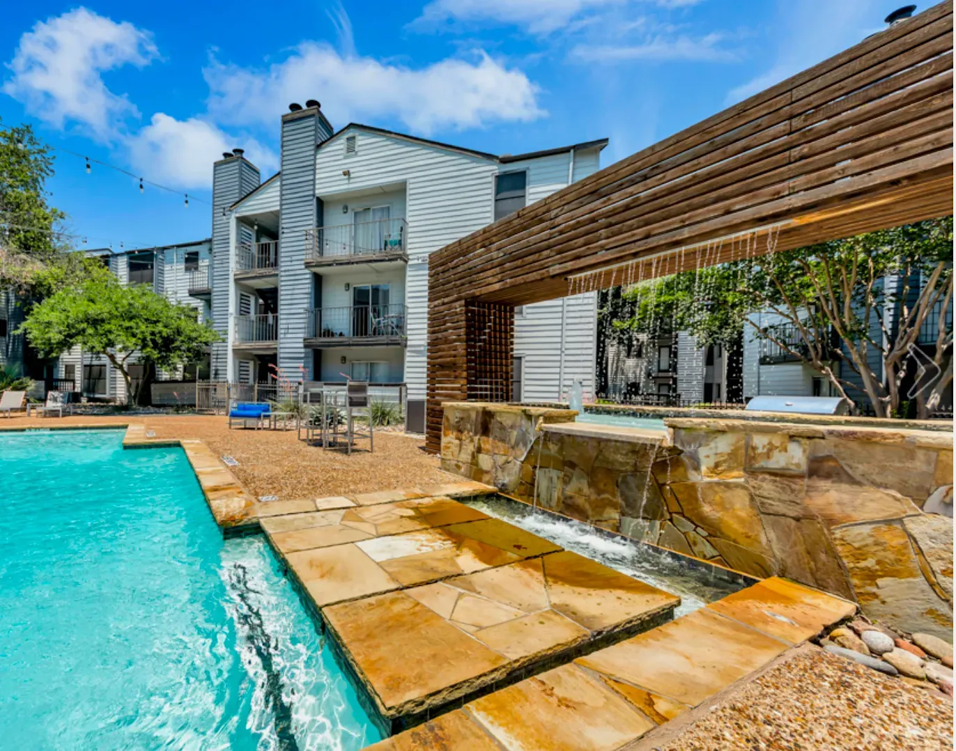 Apartment complex pool with a waterfall feature, blue water, and sunny sky.