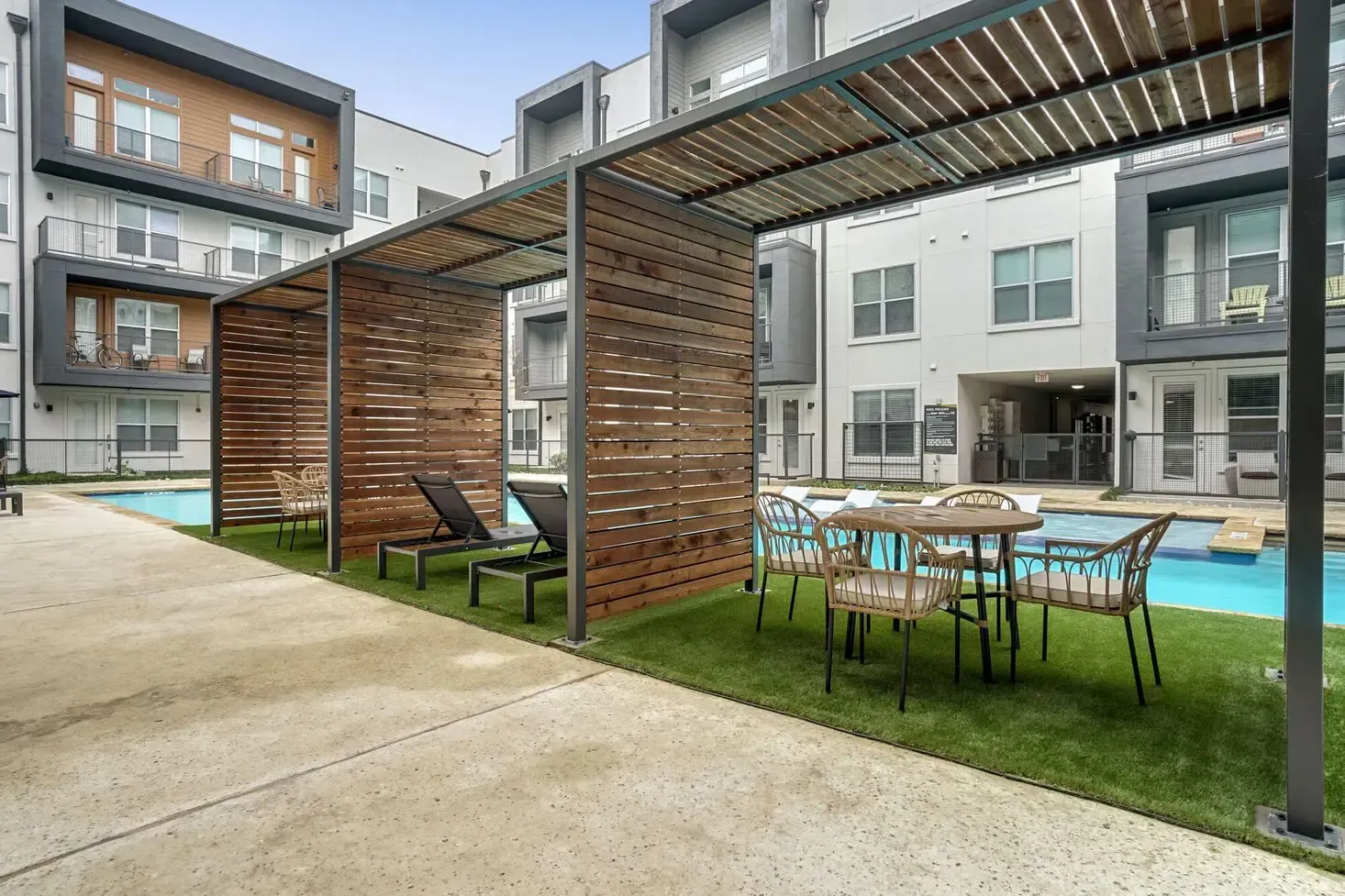 Poolside lounge area with wooden structures, seating, and an apartment building in the background.