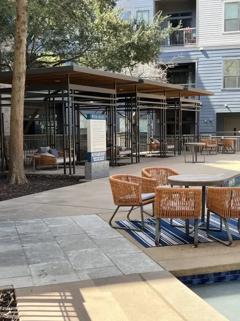 Outdoor seating area by a pool with wicker chairs, tables, and a covered structure. Blue accents.