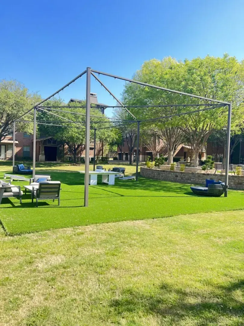 Lawn with an open metal pergola. Outdoor seating and string lights set against a brick building and trees.