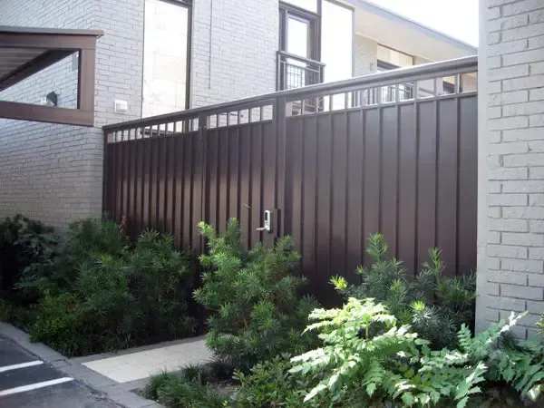 Brown metal fence with a gate, bordering greenery and a building with brick walls.