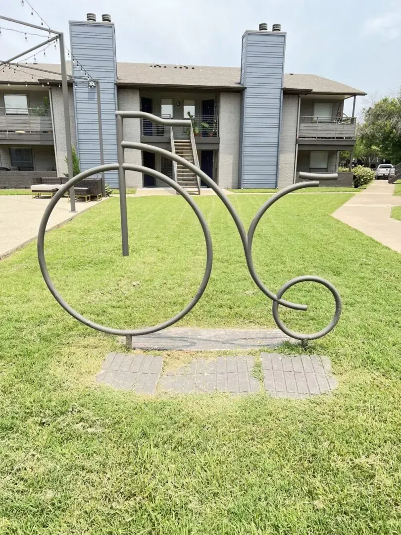 Metal sculpture of a penny-farthing bicycle on a grassy lawn, in front of a two-story apartment building.