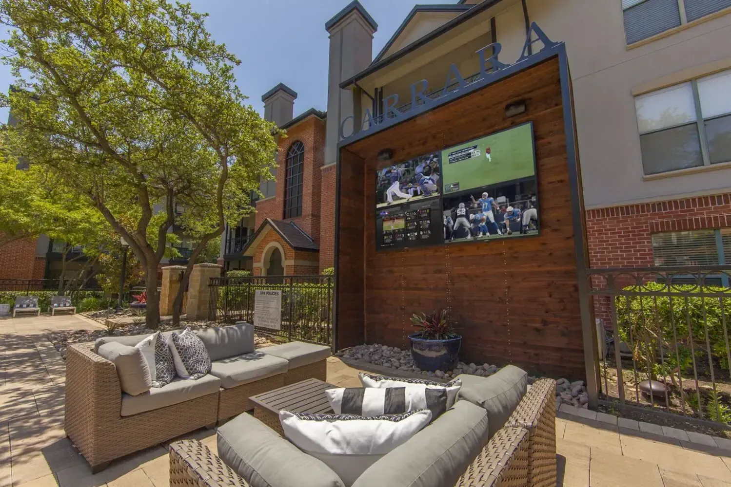 Outdoor lounge with wicker furniture, multiple TVs showing sports, and a tree.
