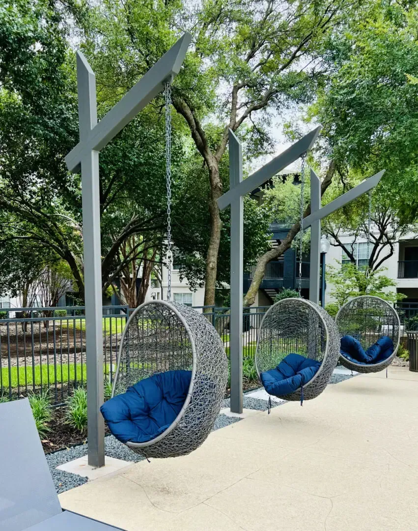 Three gray hanging chairs with blue cushions in a park setting.
