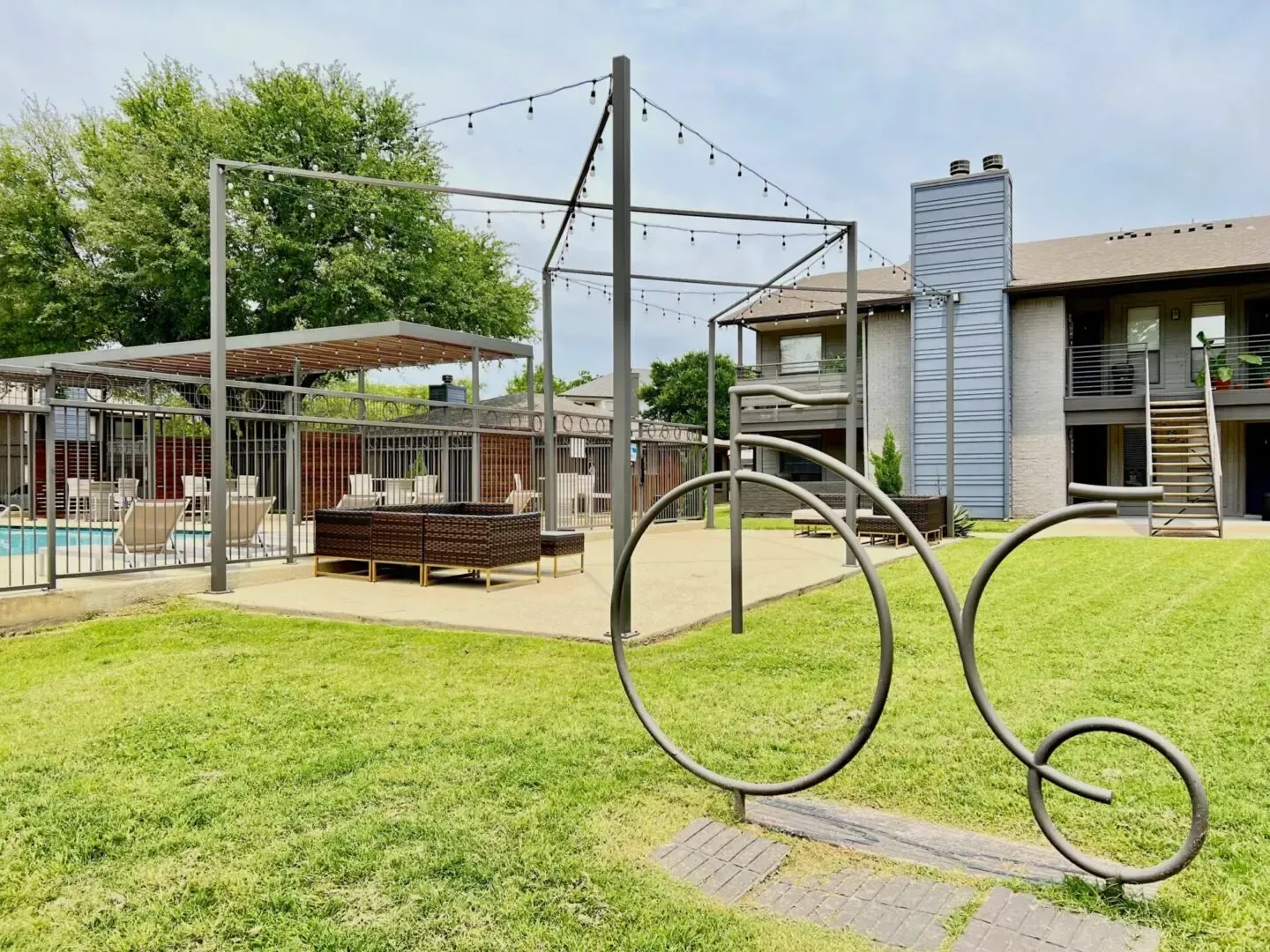Outdoor apartment seating area with metal sculptures, overhead lighting, and pool in background.