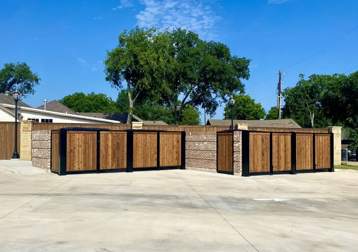 Wooden trash enclosures with black metal frames on a concrete lot, with a blue sky and trees in the background.
