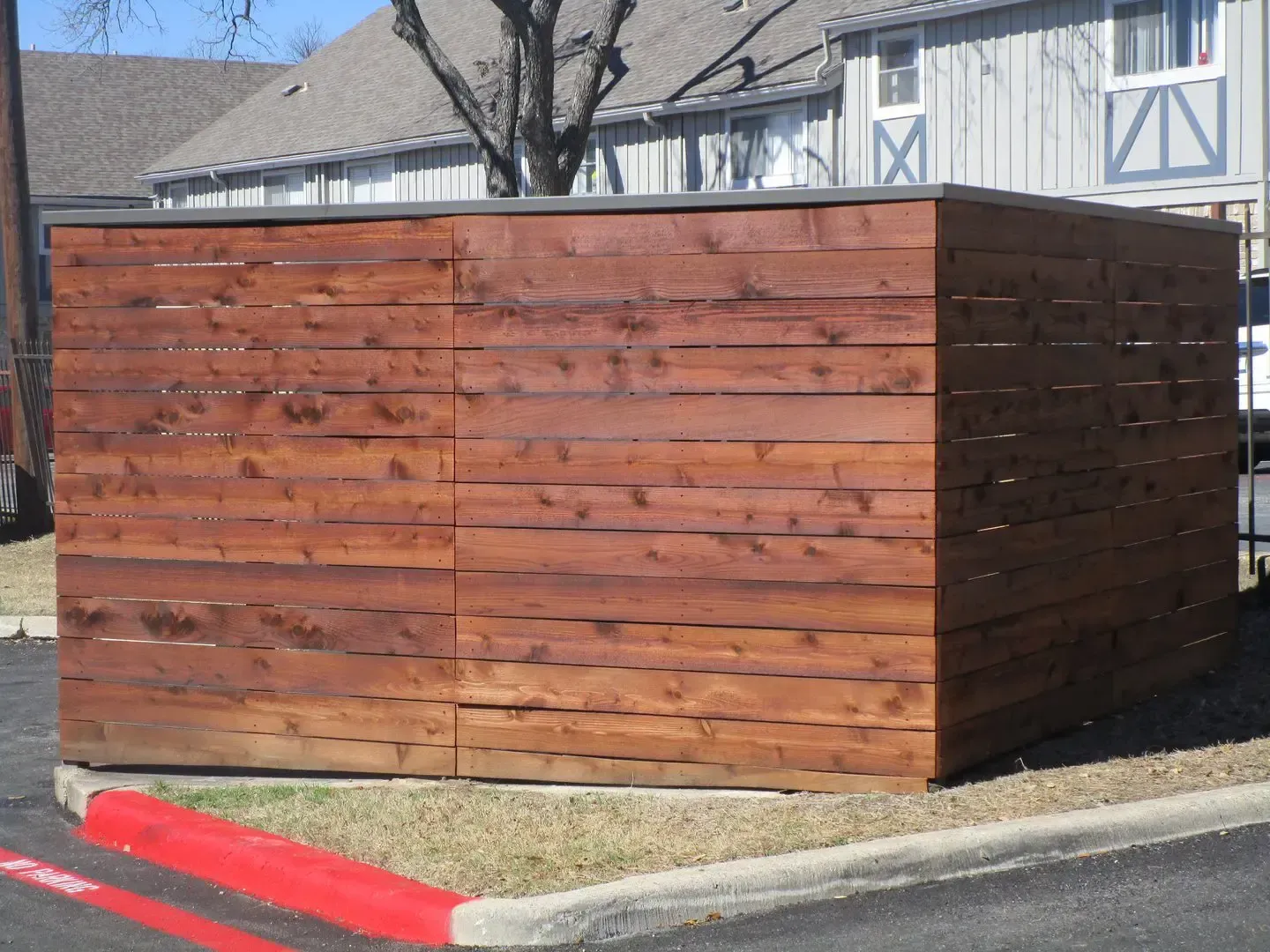 Wooden enclosure with door, in front of a building, next to a parking area with a red curb.