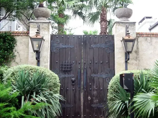 Dark wooden gate with decorative ironwork set in a stone wall, flanked by lamps and greenery.