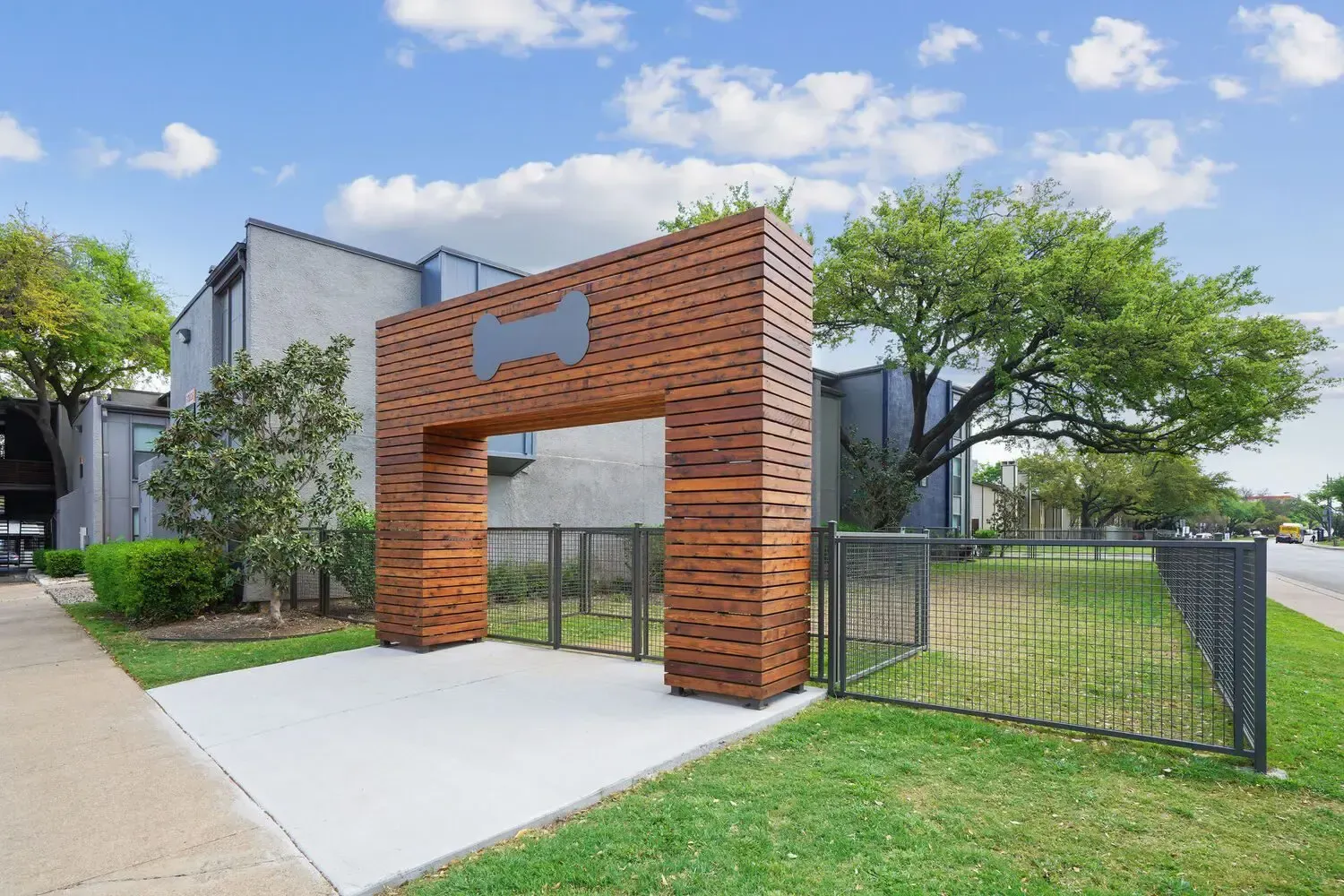 Wooden dog park entrance with a bone symbol over a concrete path and fenced area.