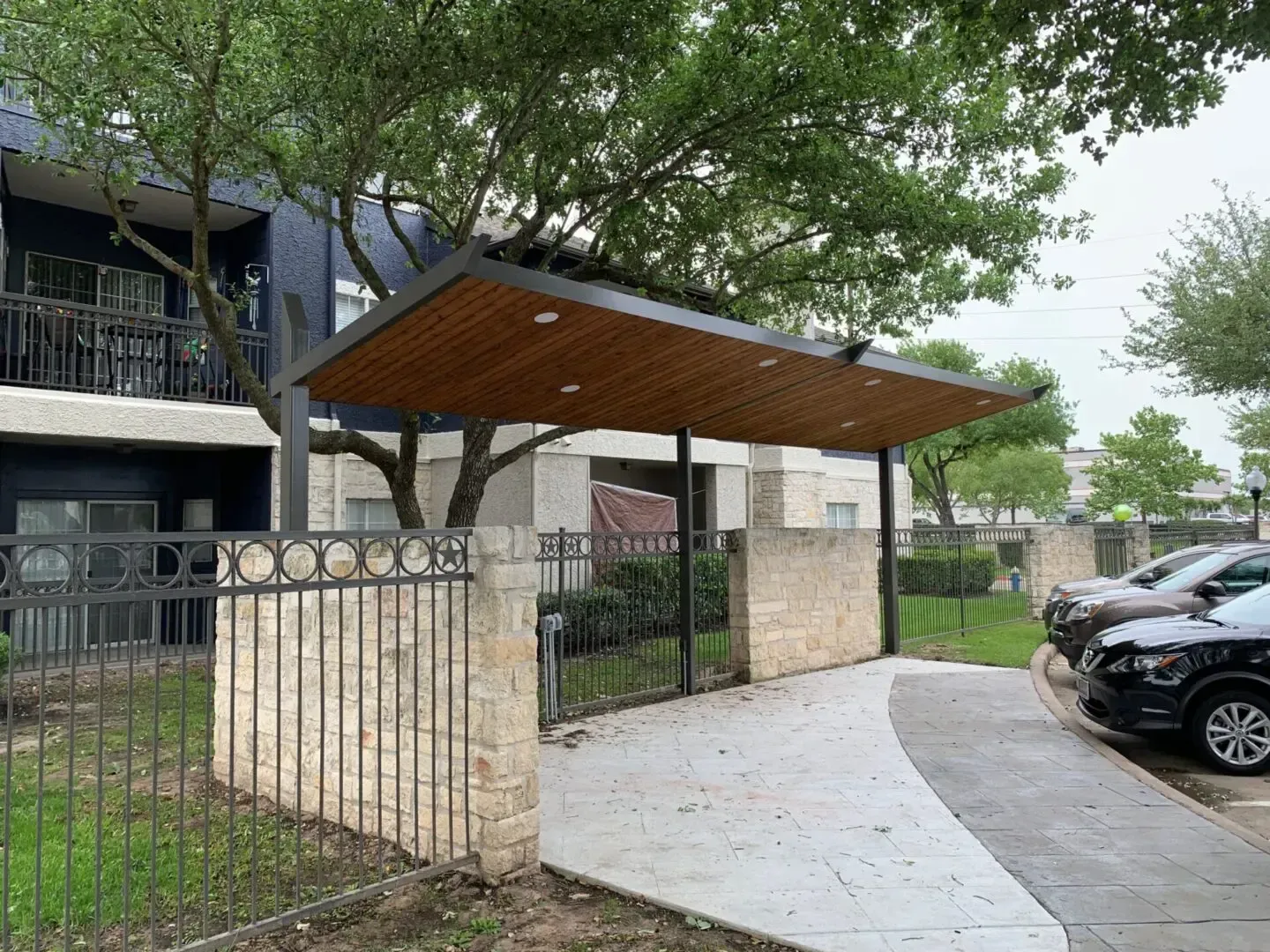 Carport entrance with wood ceiling, black supports, stone fence, gray pavement and cars.