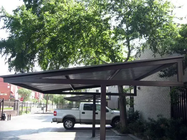 Brown metal carport over parked truck. Trees and building in background.