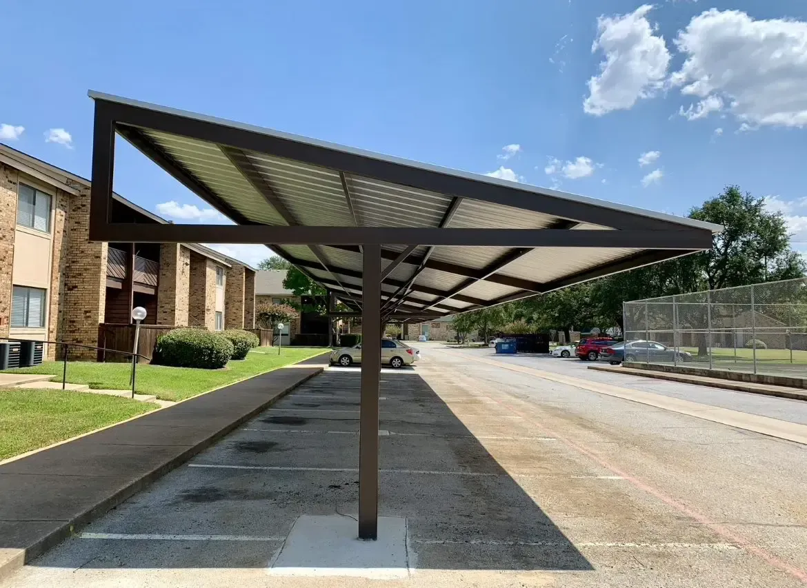 Parking lot with metal carport; brown supports, angled roof, parked cars under the shelter, apartment building in the background.