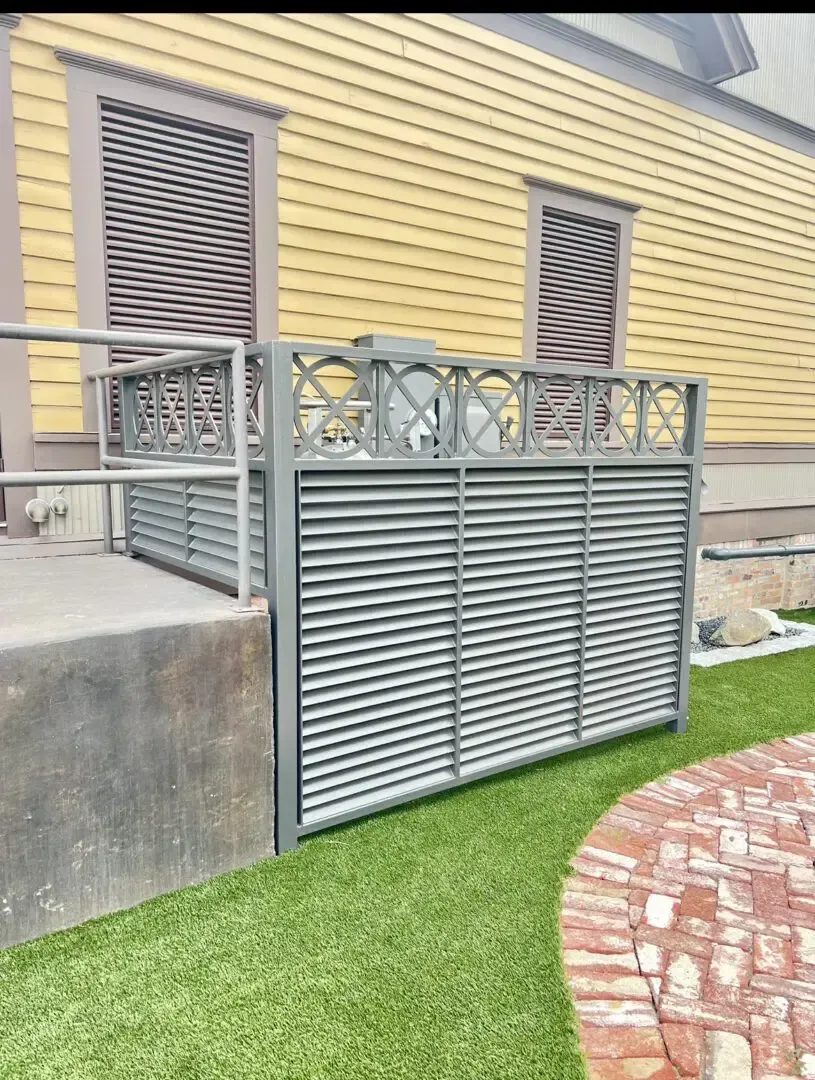 An outdoor wheelchair lift enclosed by gray louvered panels, beside a yellow building with matching window shutters.