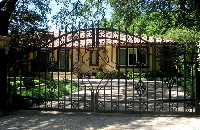 Black wrought-iron gate in front of a tan house with a driveway, trees, and landscaping.