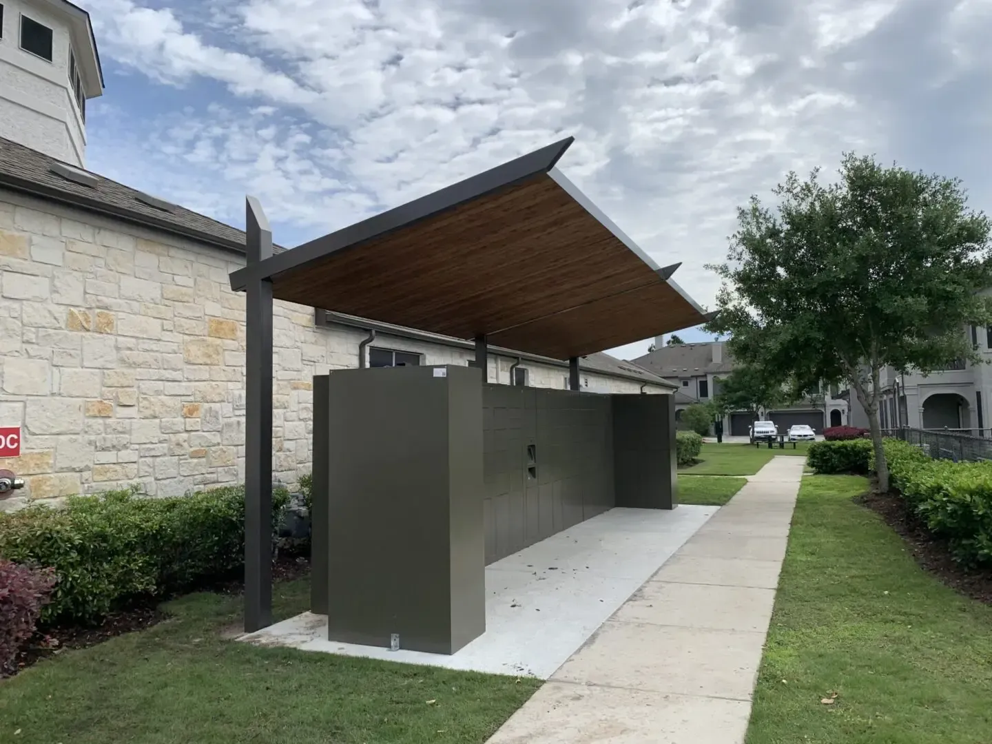 Outdoor shower station with a wooden roof, beside a building and walkway on a grassy lawn.