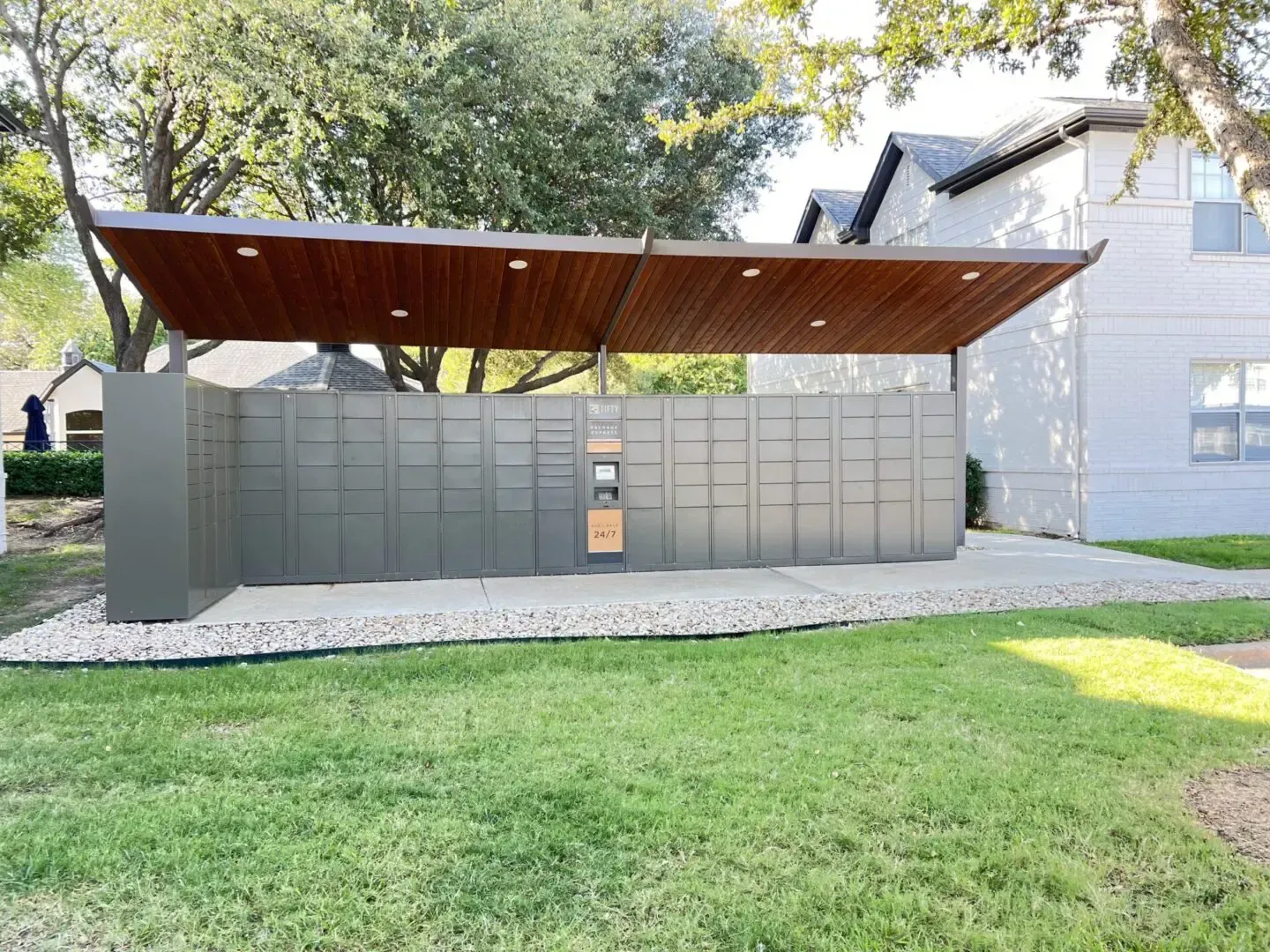 Outdoor package lockers with a wooden roof and gray metal doors, situated on a gravel bed in a grassy area.