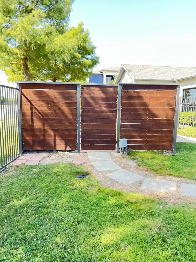 Wooden gate with dark stain, flanked by a black fence and a paved path through the grass.