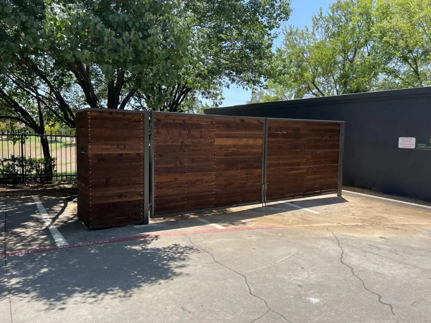 Wooden trash enclosure in a parking lot, brown wood and metal supports, a black building in the background.