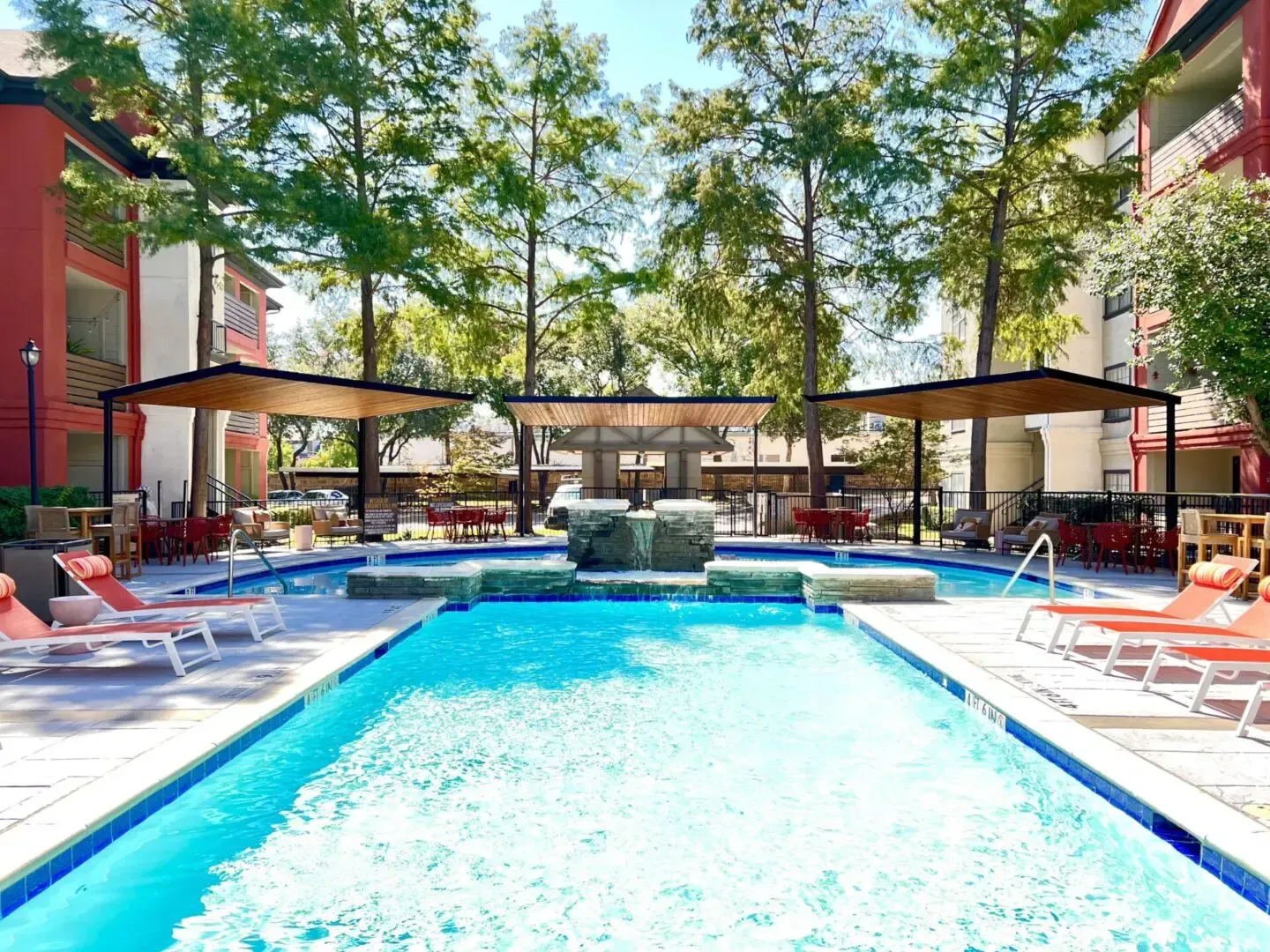Pool area with lounge chairs, shaded seating, and red apartment buildings.